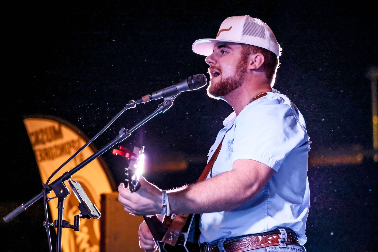 Country musician in white cap and shirt singing and playing acoustic guitar at an outdoor night concert.