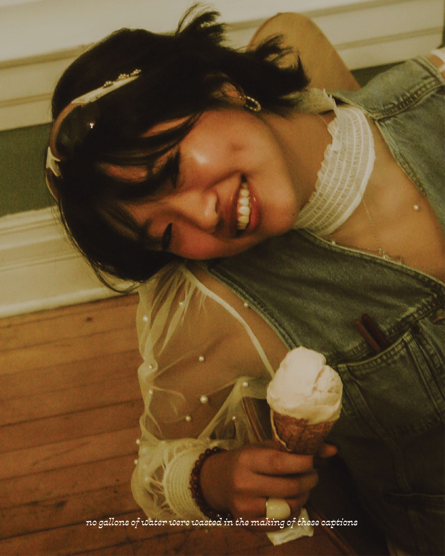 Smiling young woman holding an ice cream cone, wearing a denim vest and white lace top, leaning forward on a wooden floor.