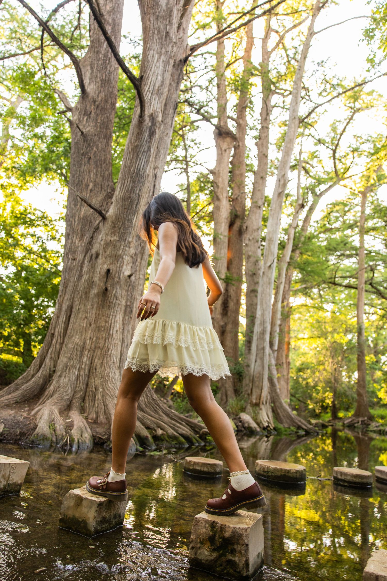 Young girl in white dress stepping on stone path through a lush green forest near water.