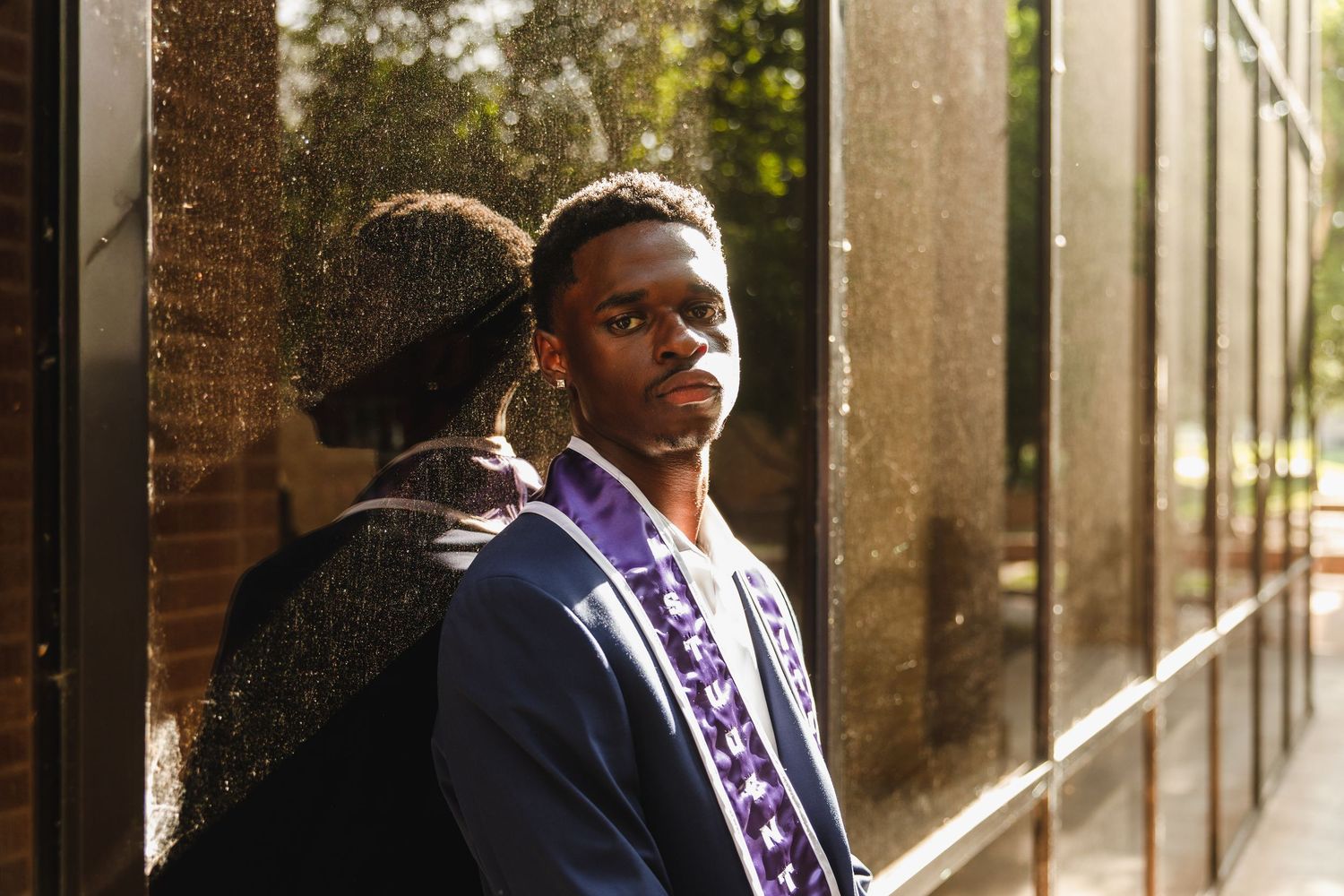 Young man in graduation stole leans against glass building reflecting his image outdoors.