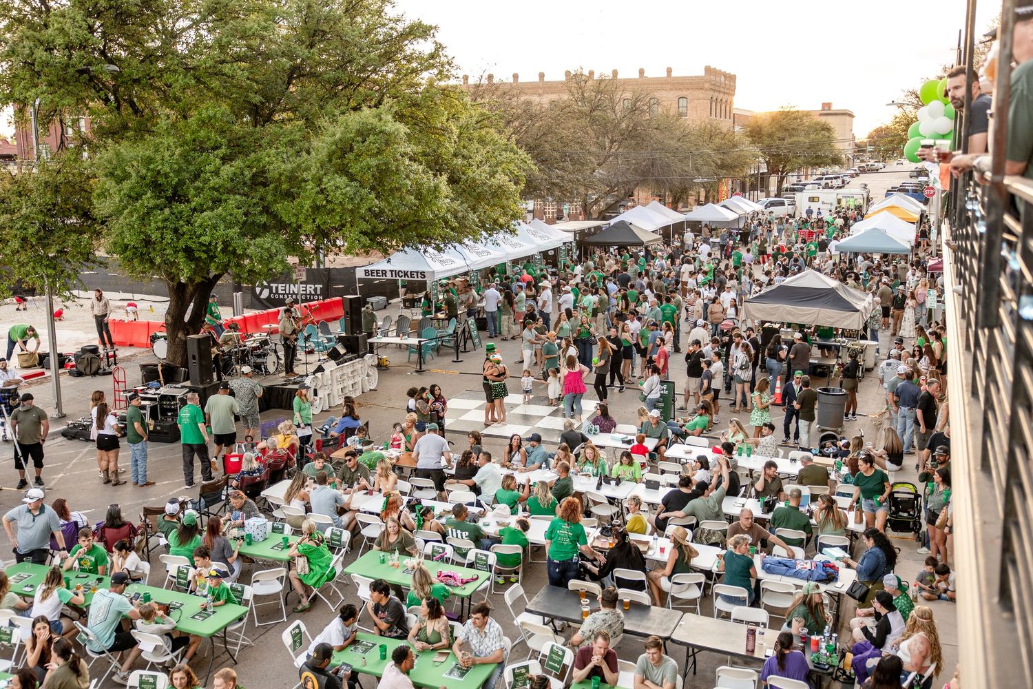 Aerial view of a large outdoor festival with crowds in green attire at street tables.