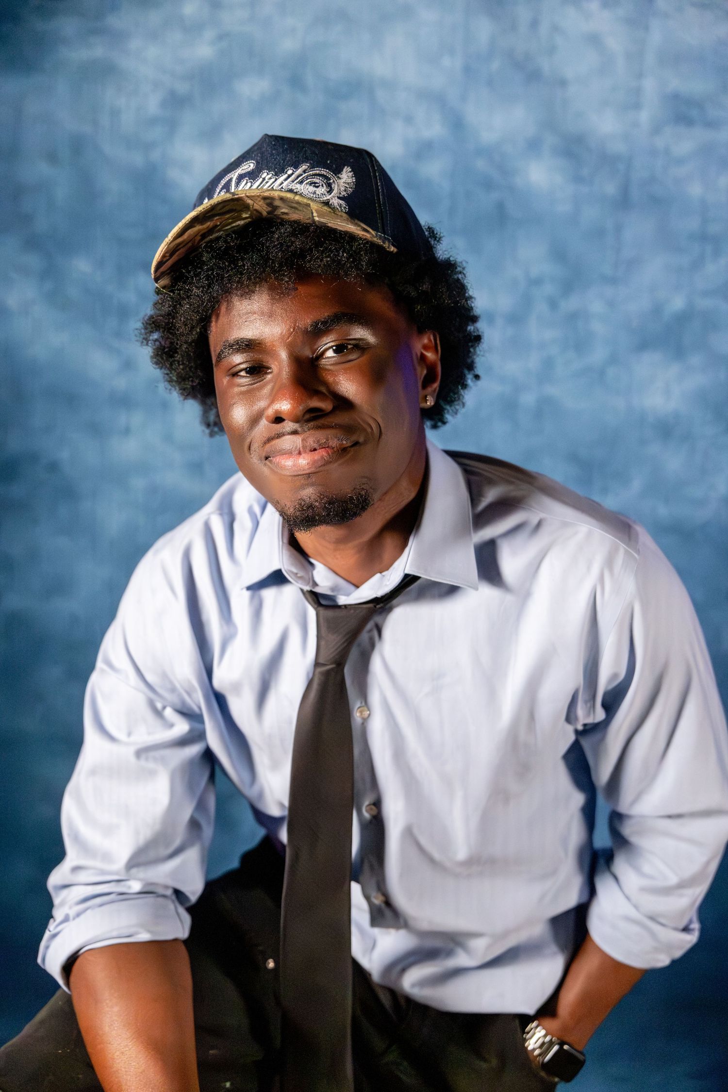 Smiling young man in a navy cap and tie poses against a blue studio background.