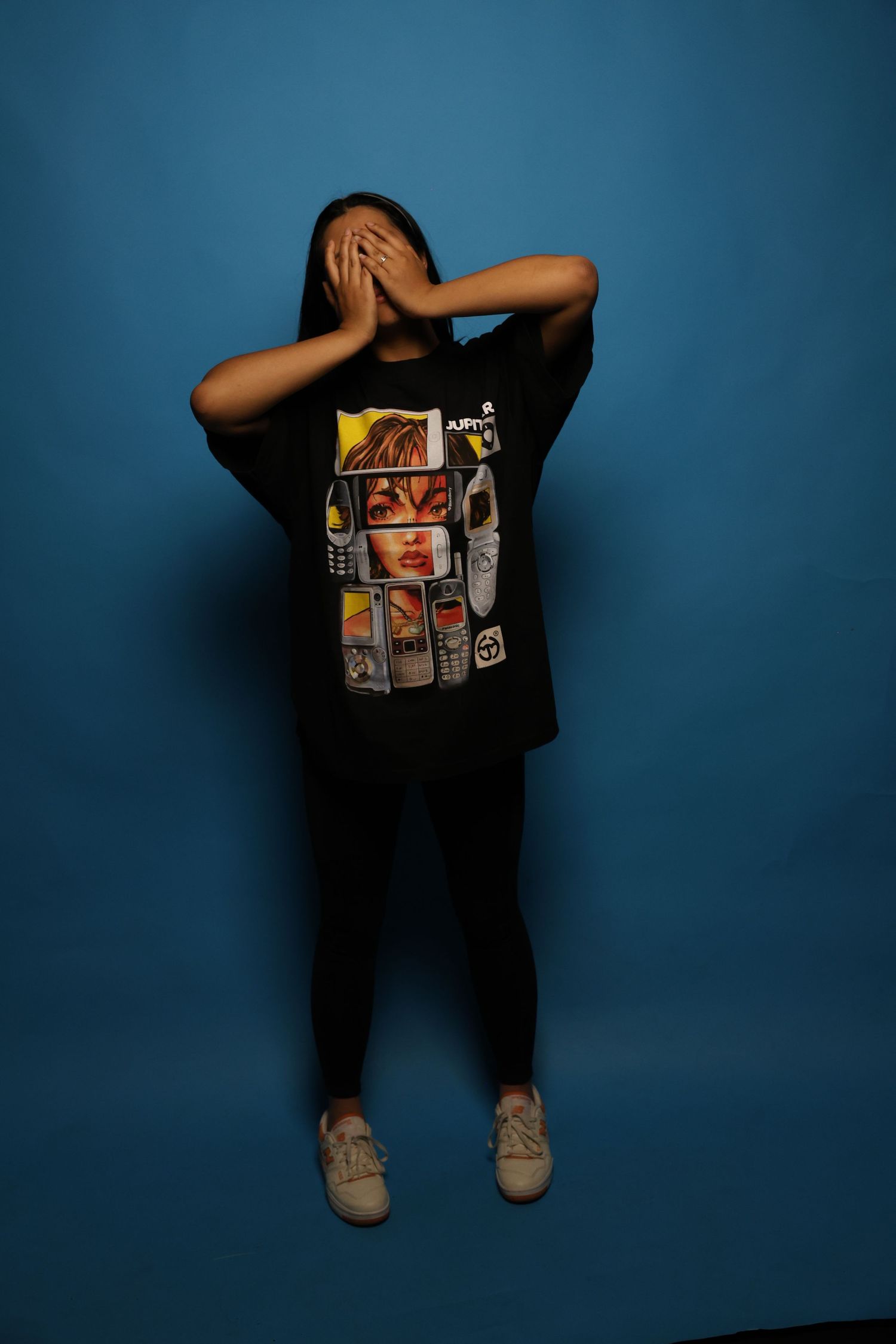 Person in oversized black Jupittar graphic tee covers face with both hands on blue backdrop.