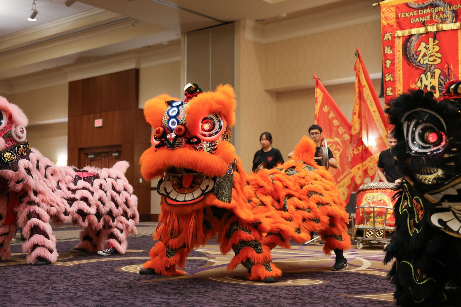 Three vibrant Chinese lion dance costumes in pink, orange, and black perform together in a decorated ballroom.