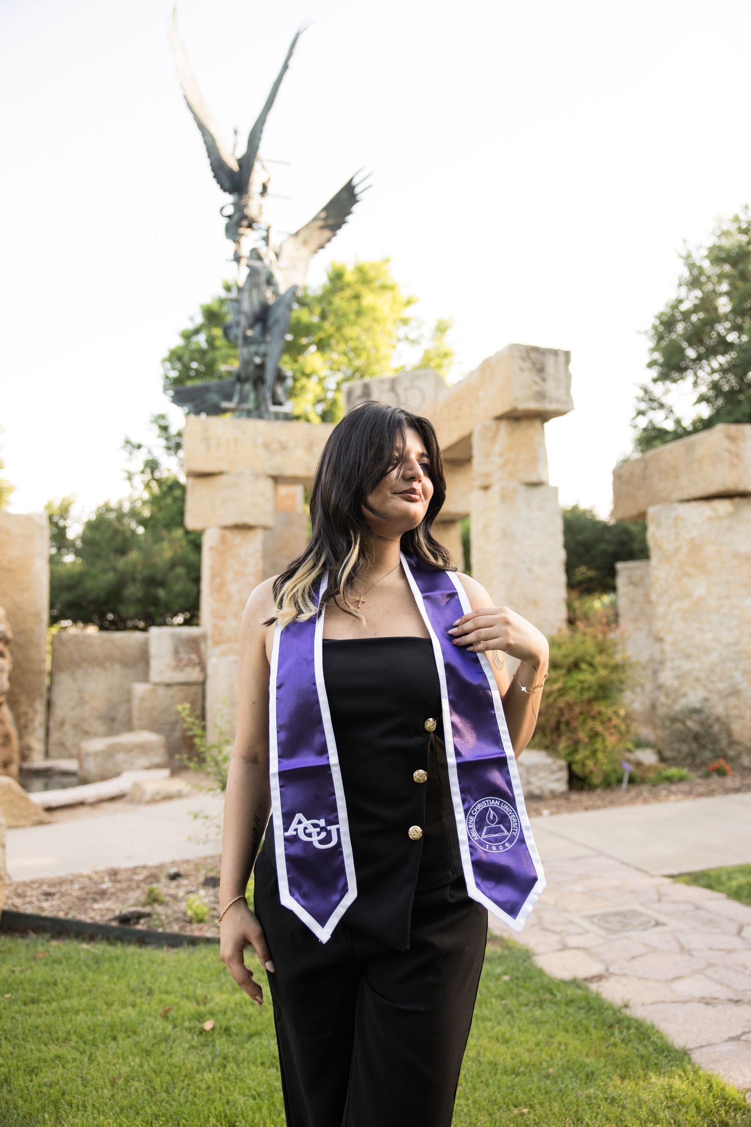 Female graduate wearing ACU purple stole posing near eagle statue on sunny campus grounds.
