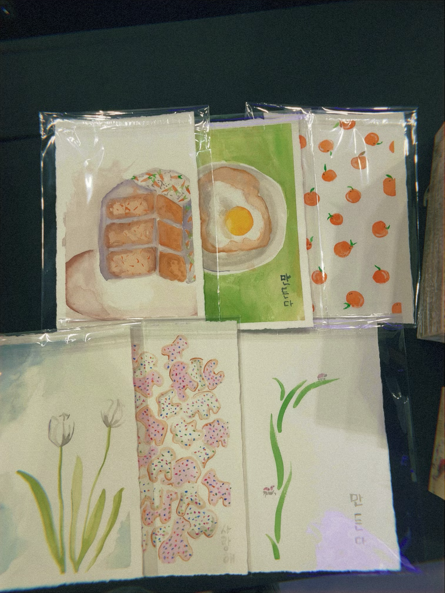Five handmade watercolor greeting cards featuring food, florals, and botanicals displayed on a dark surface.