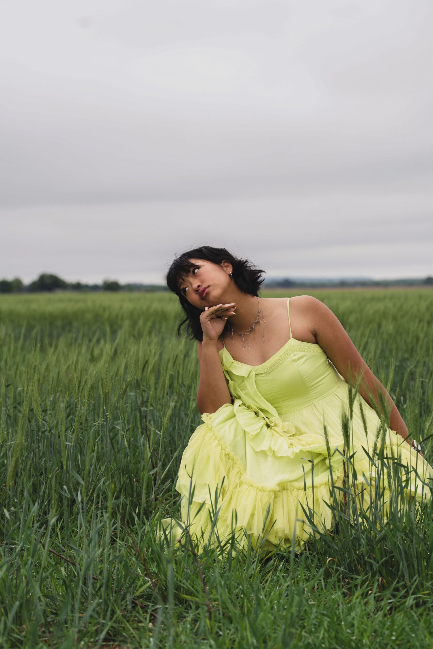 A woman in a yellow-green ruffled dress sits gracefully in a lush green field under an overcast sky.