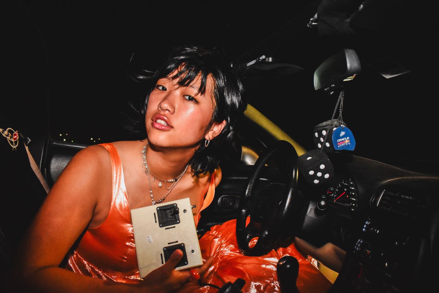 Young woman in orange satin dress sits in car holding a Polaroid camera at night.