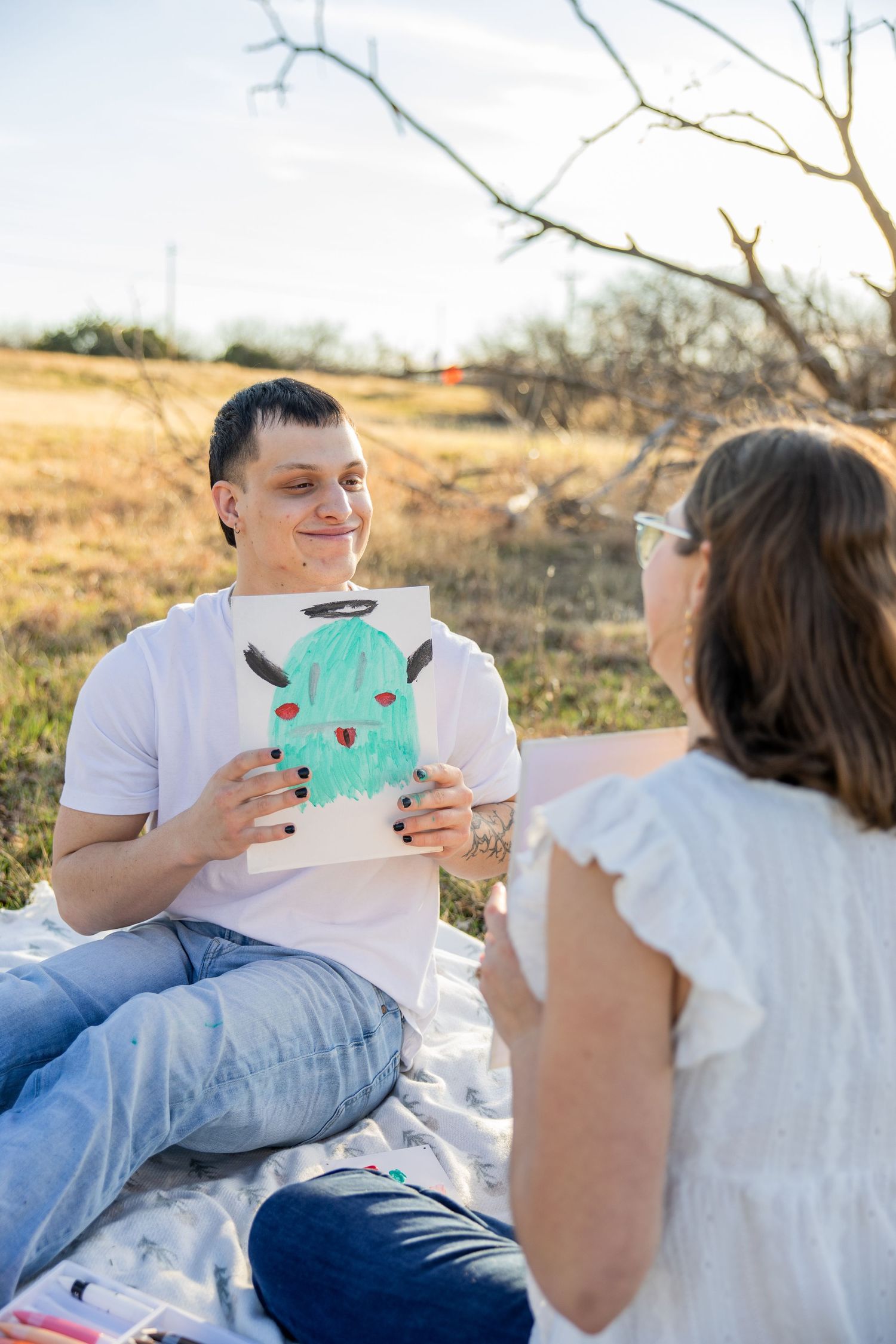 Smiling man holding up a colorful painting of a cactus during an outdoor picnic date.