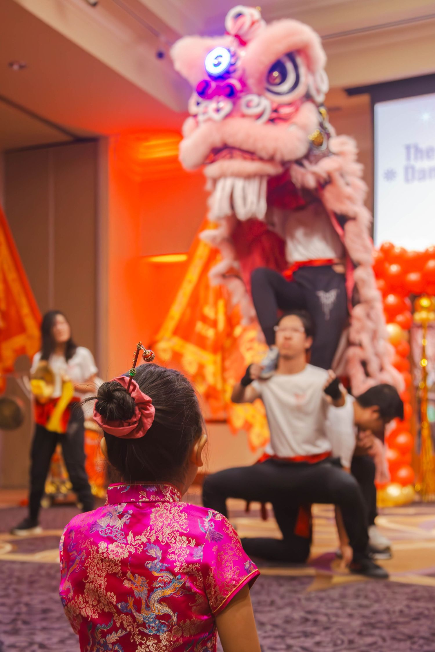 A girl in a pink qipao watches an illuminated lion dance performance at a Lunar New Year celebration.