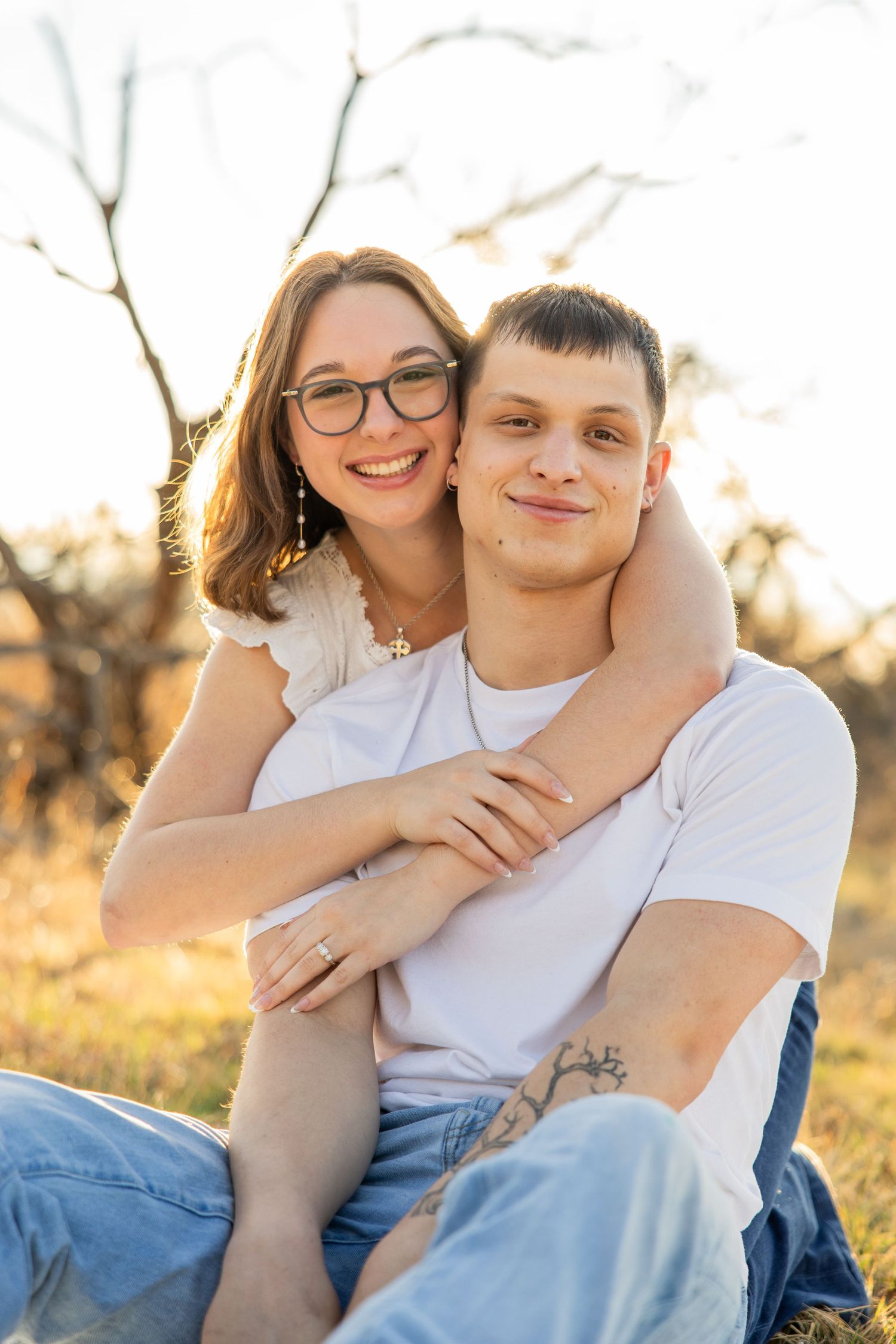 Smiling couple sitting together in a sunny field, woman with glasses embracing man in white shirt.