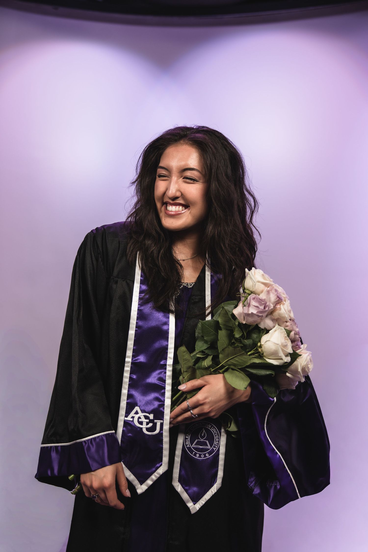 Smiling female graduate in black gown with ACU purple stole holding bouquet of flowers.