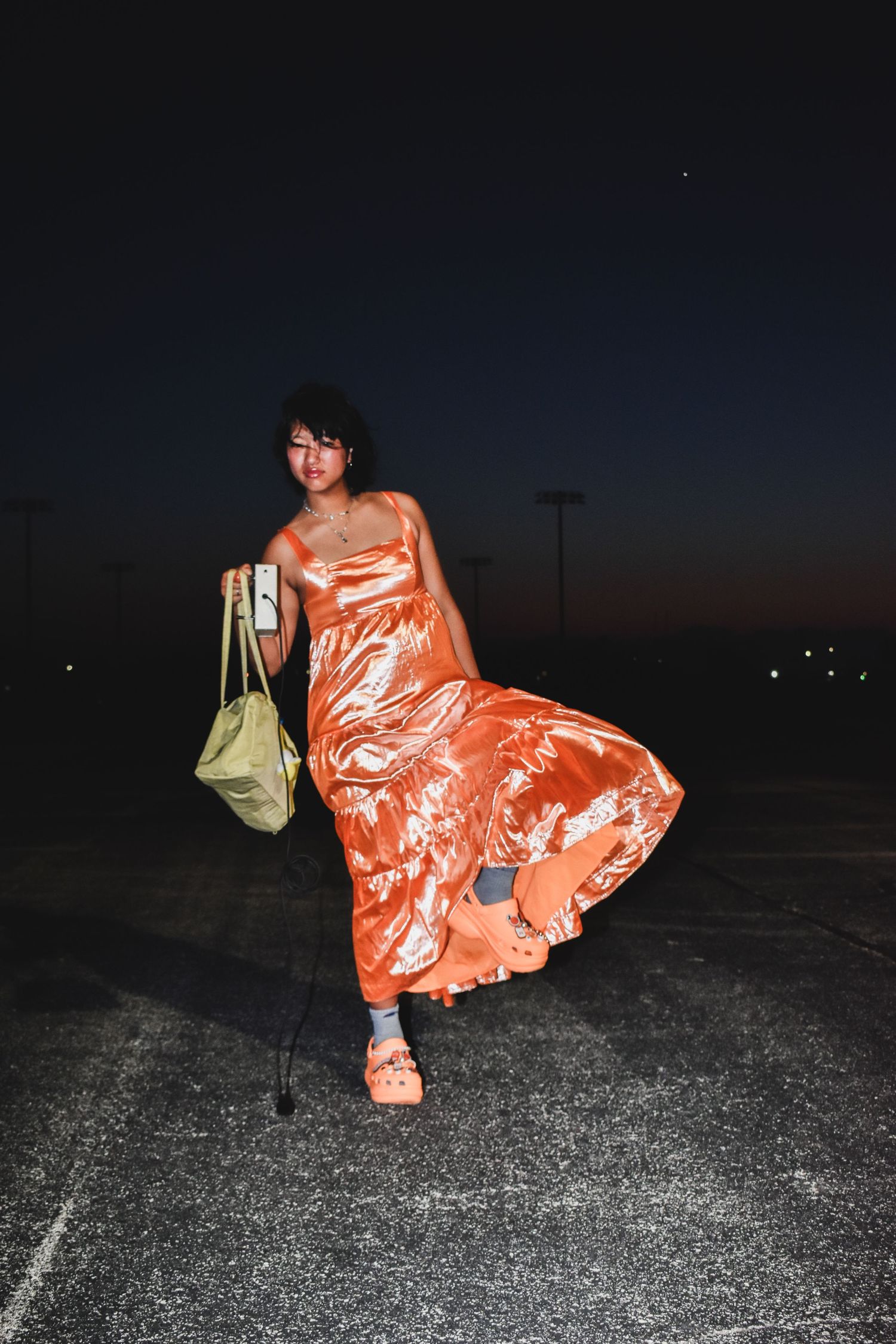 Woman in flowing orange metallic dress posing in parking lot at night with green bag.
