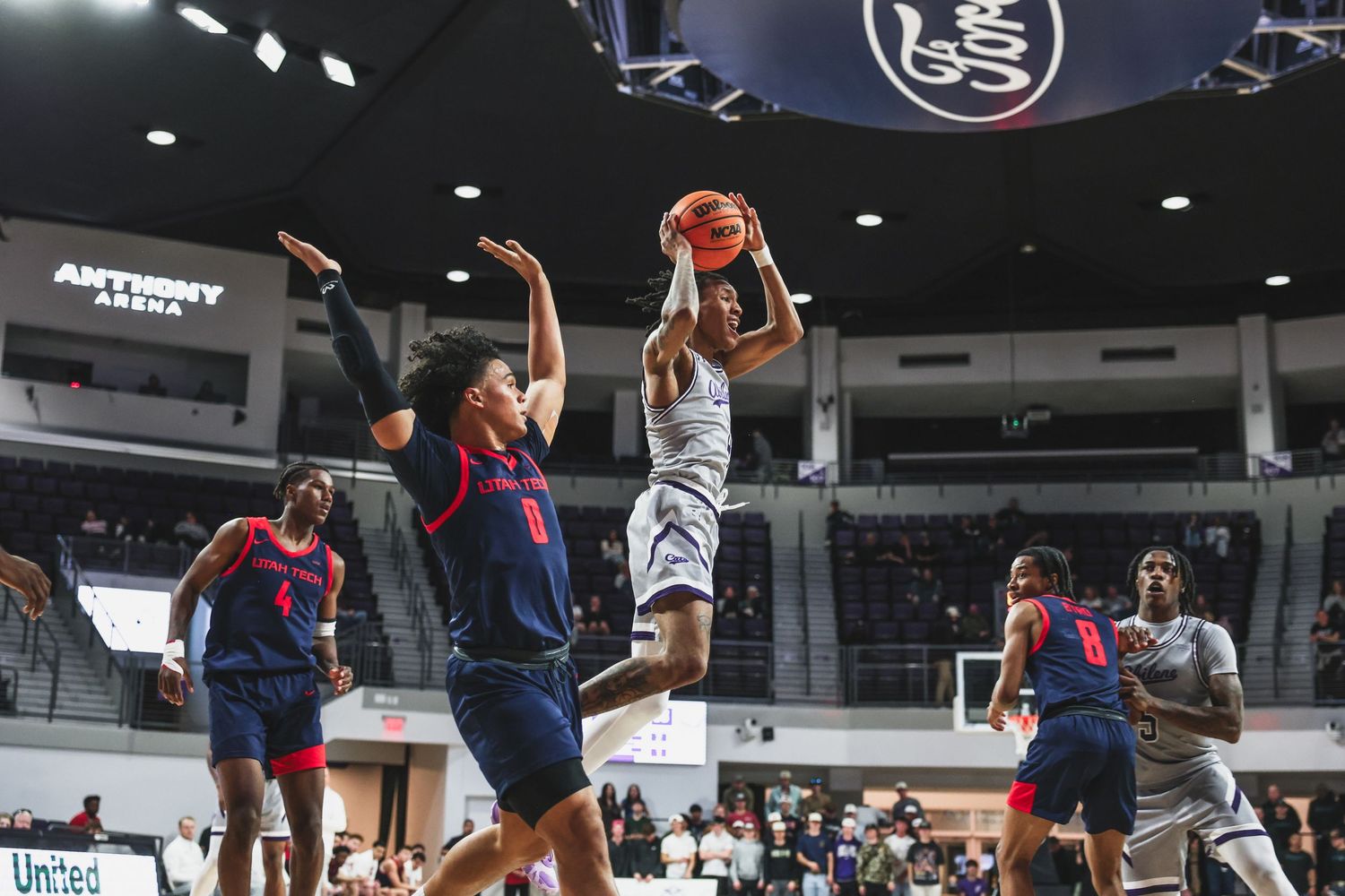 Basketball player drives to the basket for a layup against defenders in a college arena game.