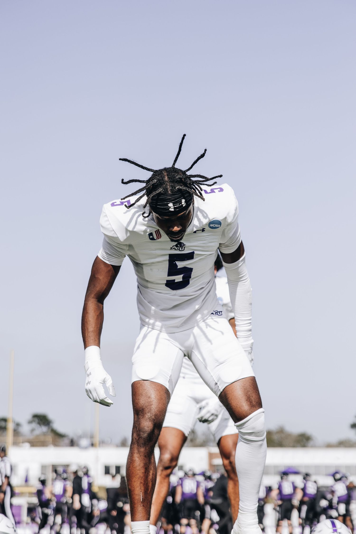 ACU football player #5 with dreadlocks leans forward intensely during a sunny game day.
