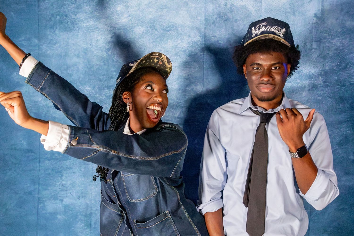 Two young people in navy caps laugh and pose energetically against a blue backdrop.