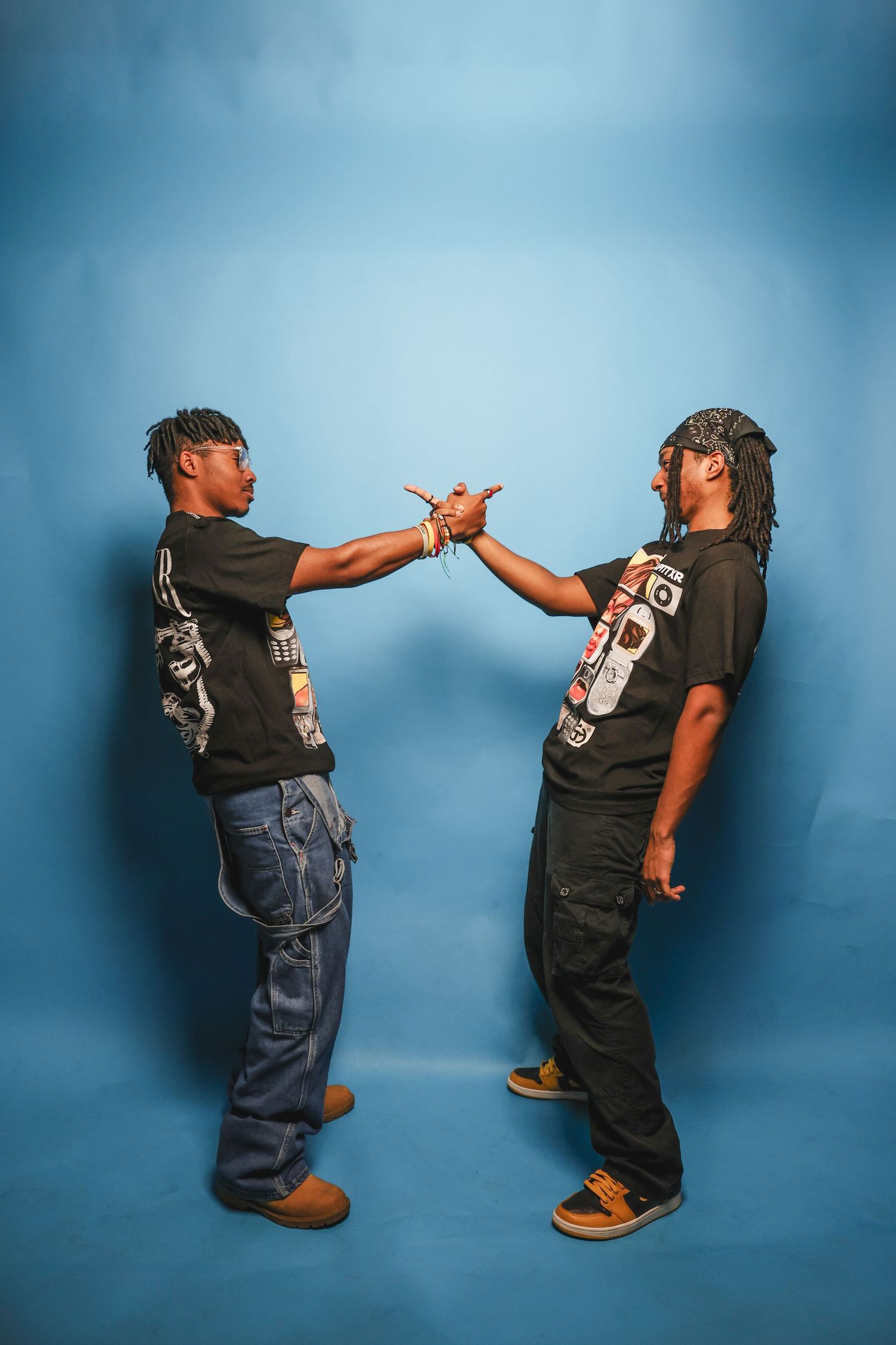 Two men in matching black graphic tees face each other doing a handshake against blue background.