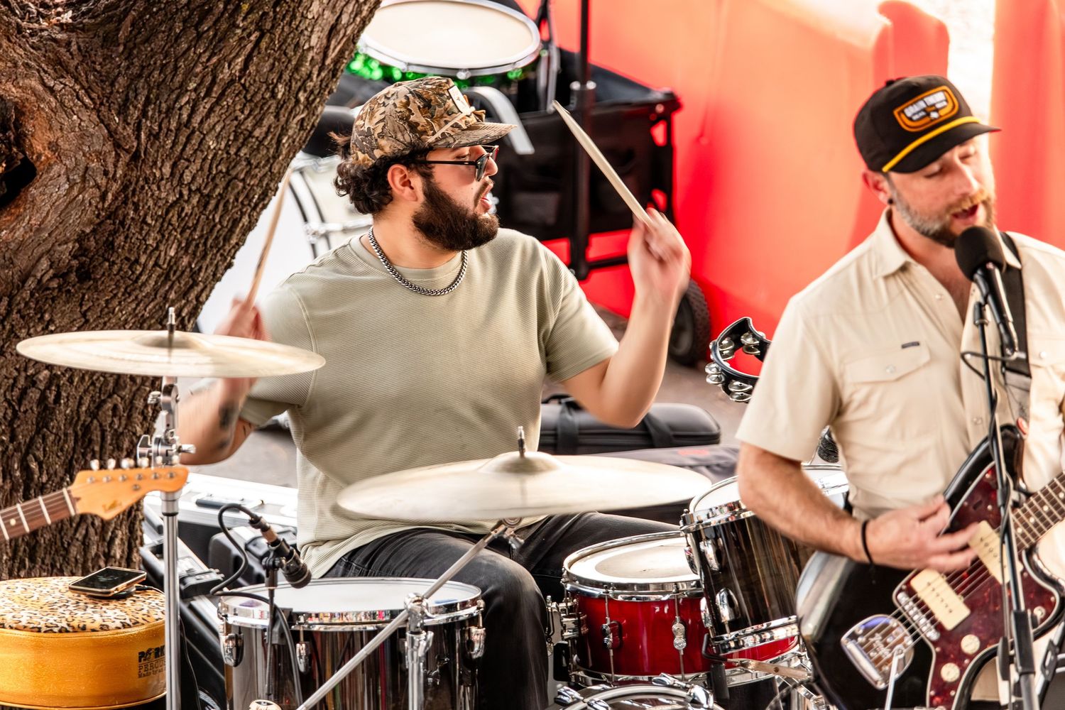 Two musicians perform outdoors, one playing drums and another playing guitar at a live music event.