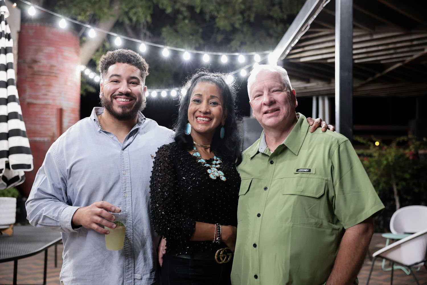 Three smiling guests pose together under string lights at an outdoor evening social gathering.