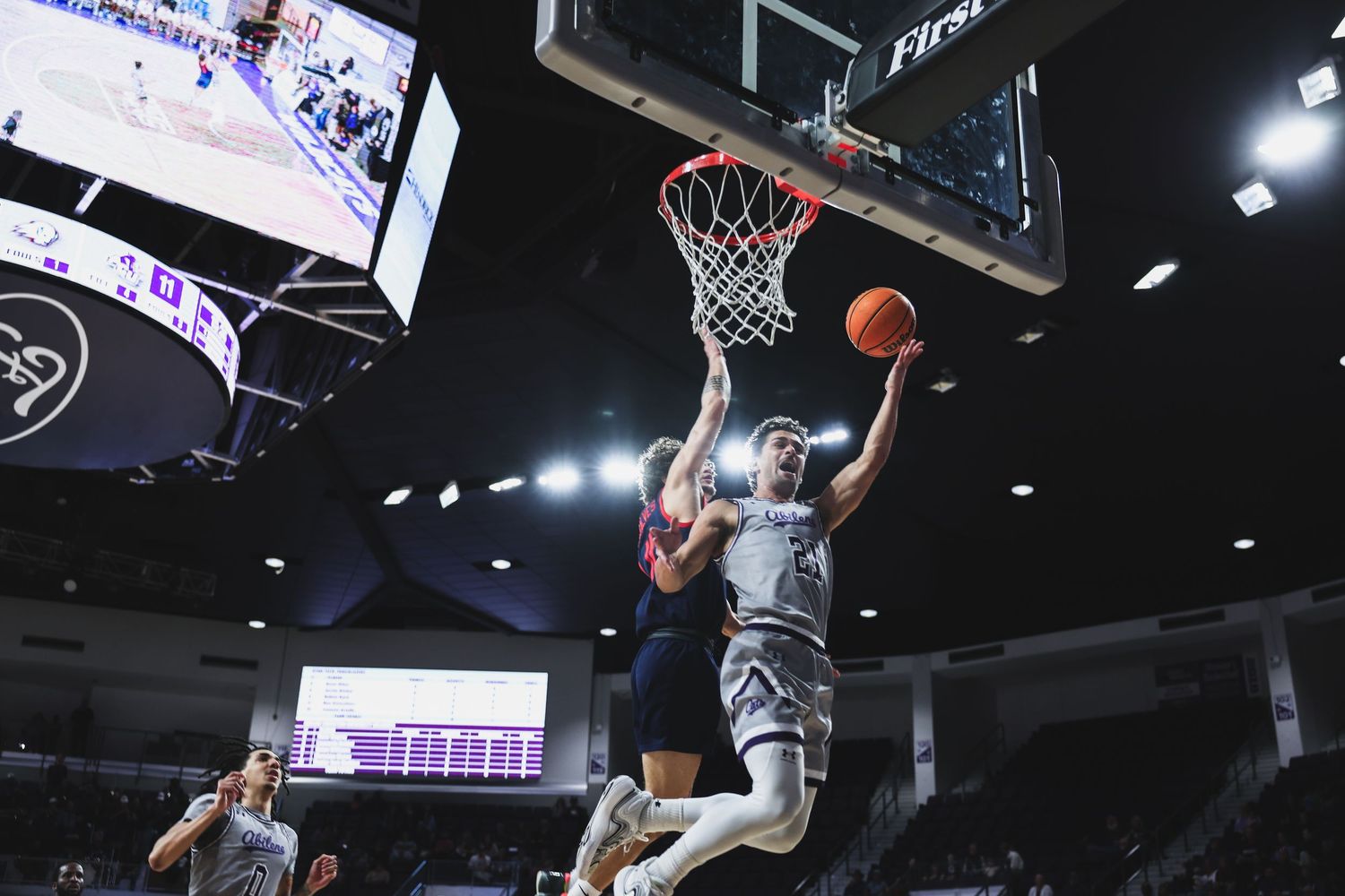 College basketball player rises for a layup past defenders under the hoop in a well-lit indoor arena.