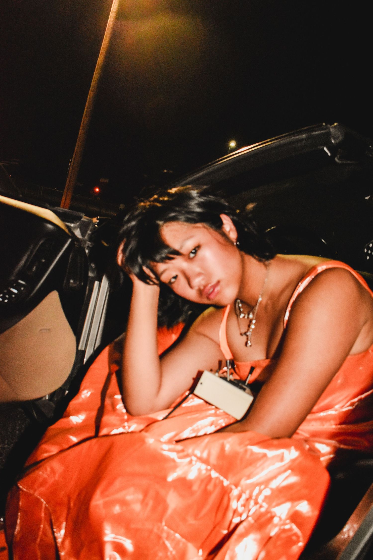 A young woman in a shiny orange dress leans against a car door at night, holding a small clutch purse.