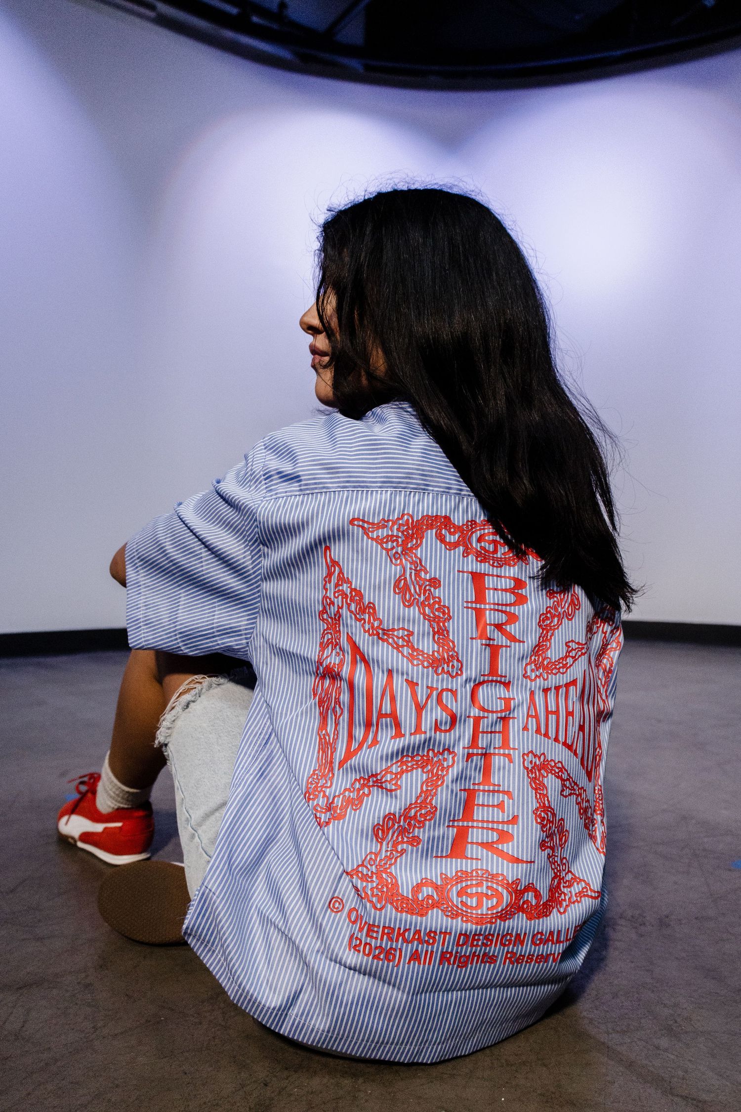Woman sits on floor showing back of blue striped shirt with red Days Brighter graphic print.