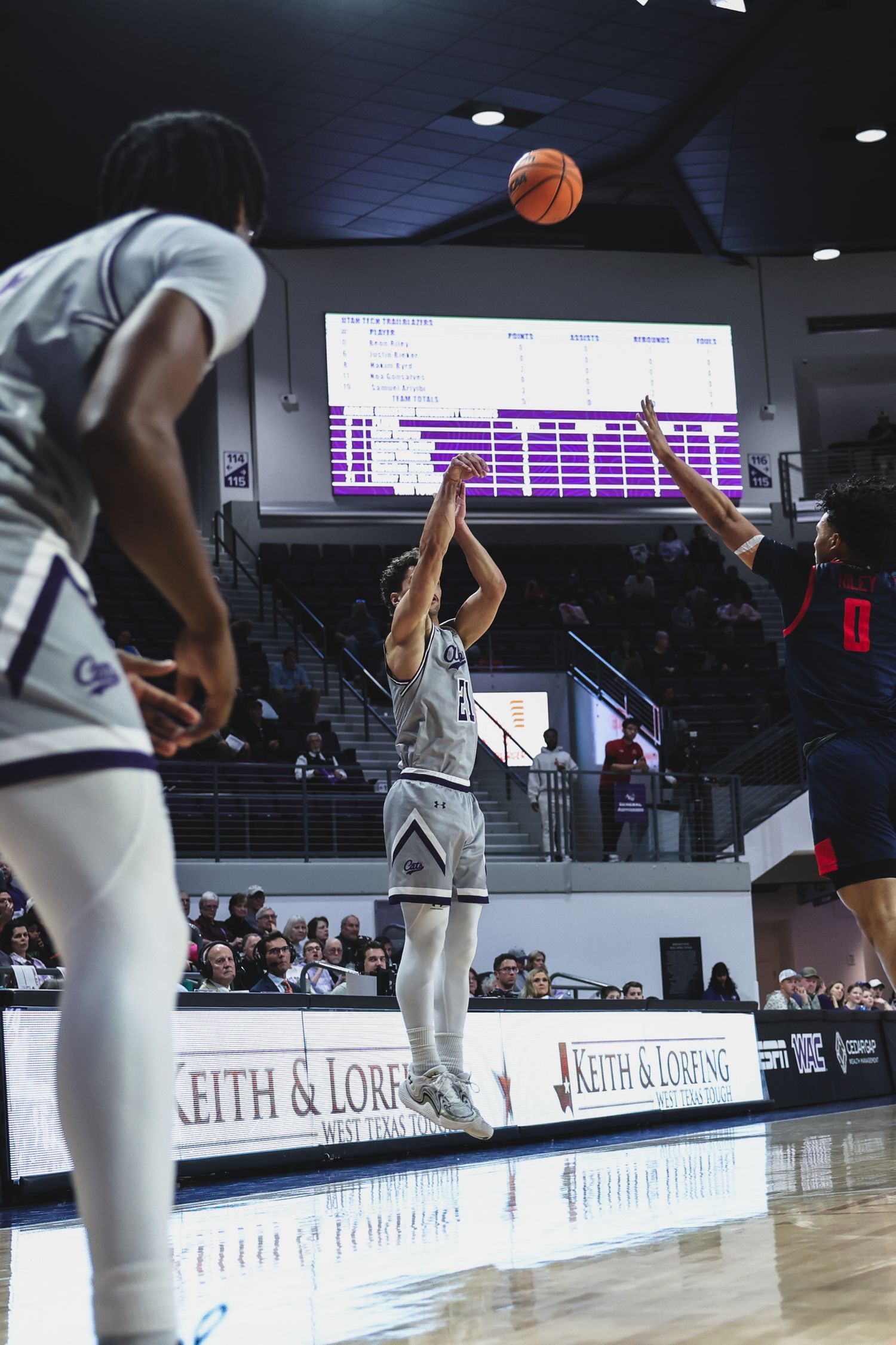 College basketball player releases a jump shot while a defender contests the shot in an indoor arena.
