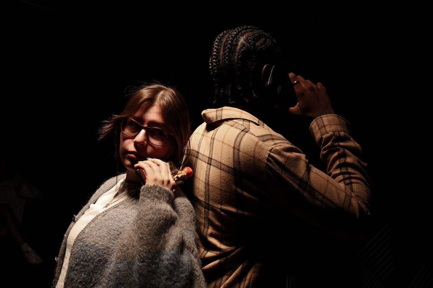 Two people in a dramatic dark scene, one in glasses and gray sweater, other in plaid jacket.