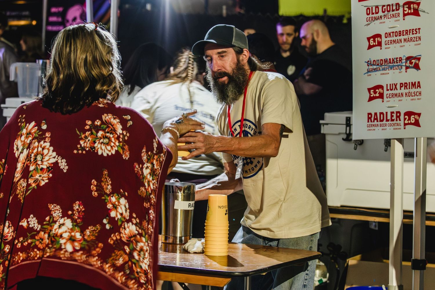 Bearded man serving drinks to a customer at an indoor Oktoberfest event at night.
