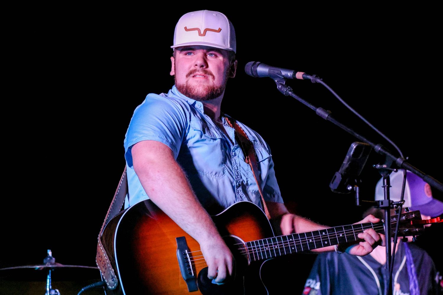 Country musician playing acoustic guitar and singing into microphone on stage under warm lights.