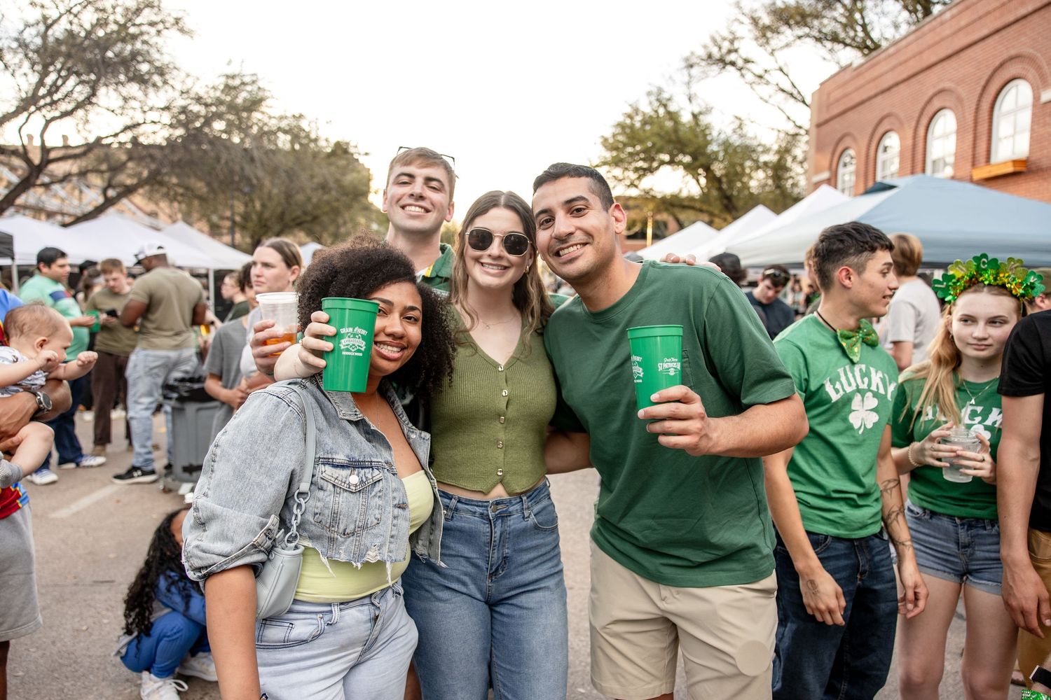 Group of smiling friends pose together holding green cups at a St. Patricks Day street festival.