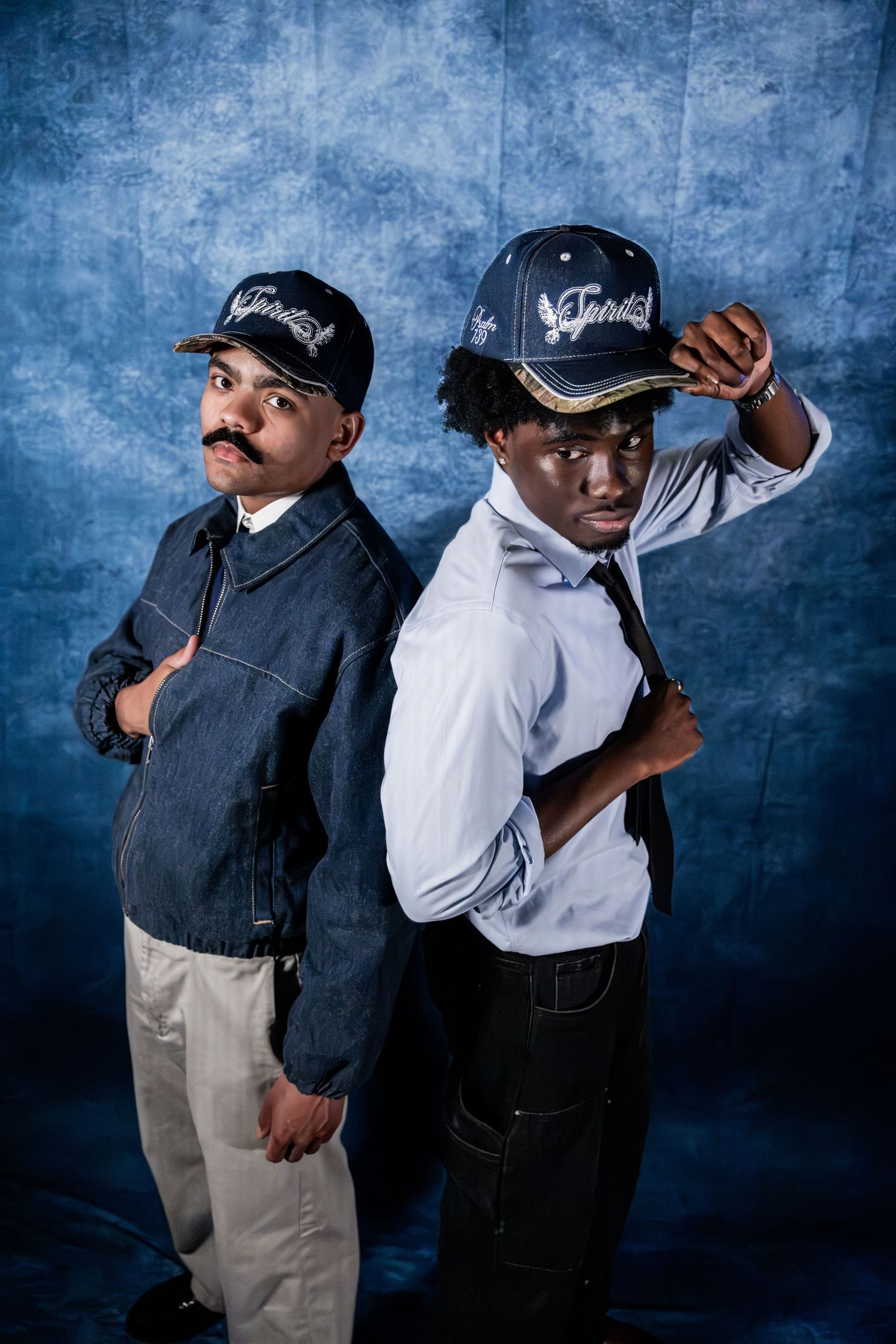 Two young men in navy caps pose back-to-back against a blue textured studio background.