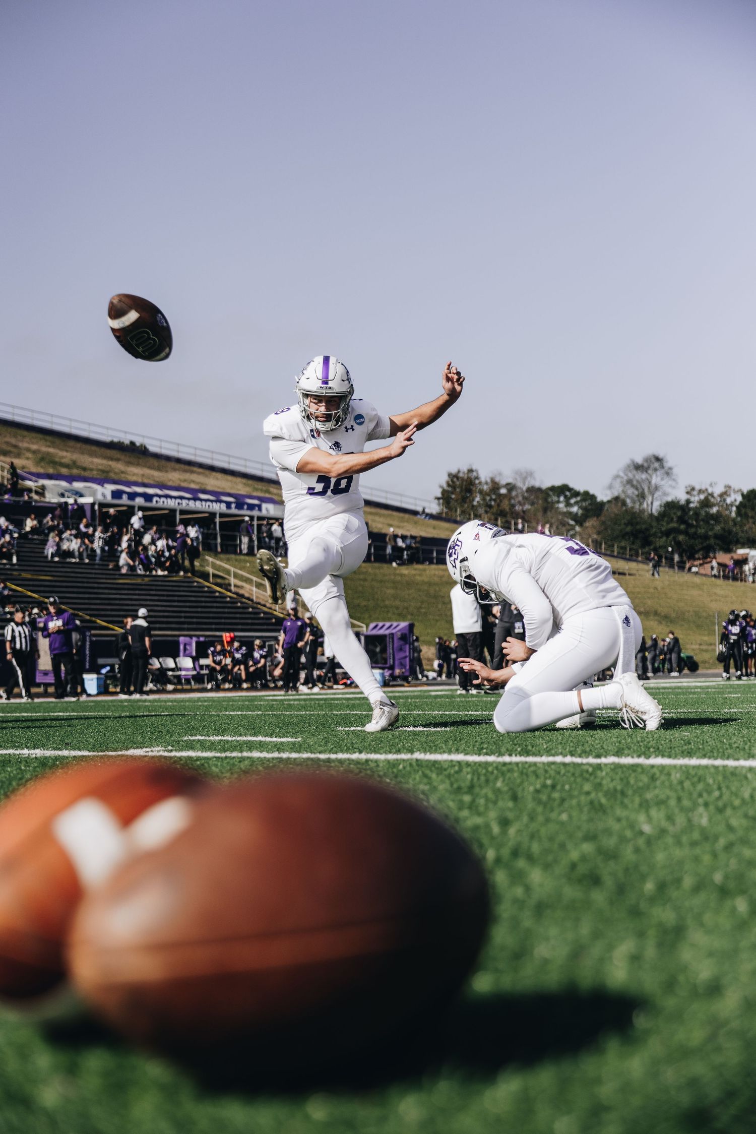 College football player executing a field goal kick during a sunny outdoor stadium game.