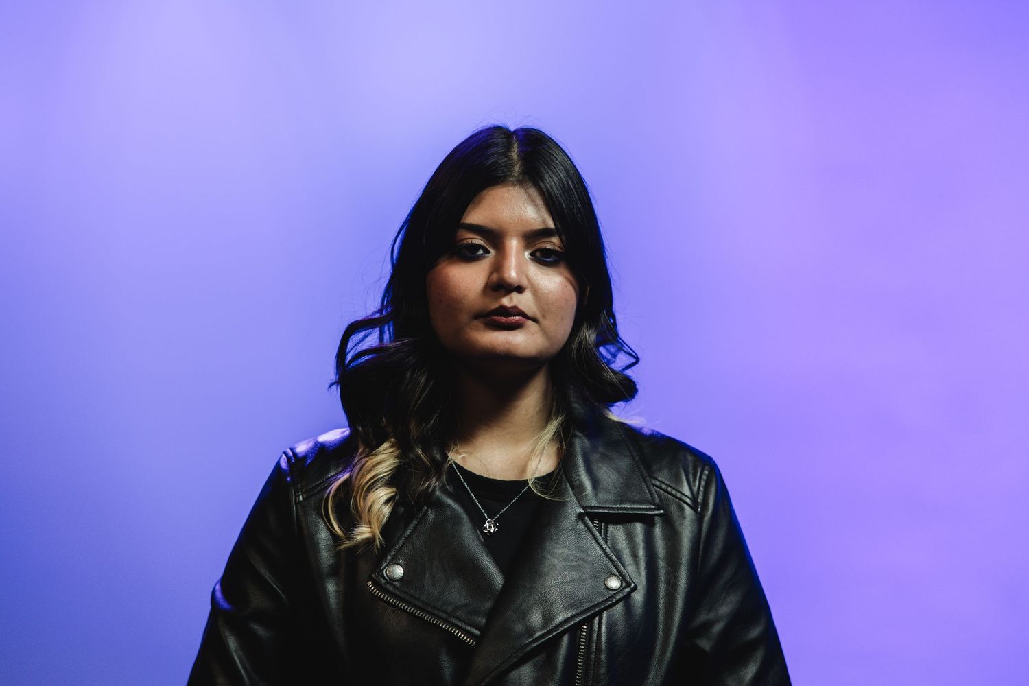 Young woman in black leather jacket against purple background with serious expression.