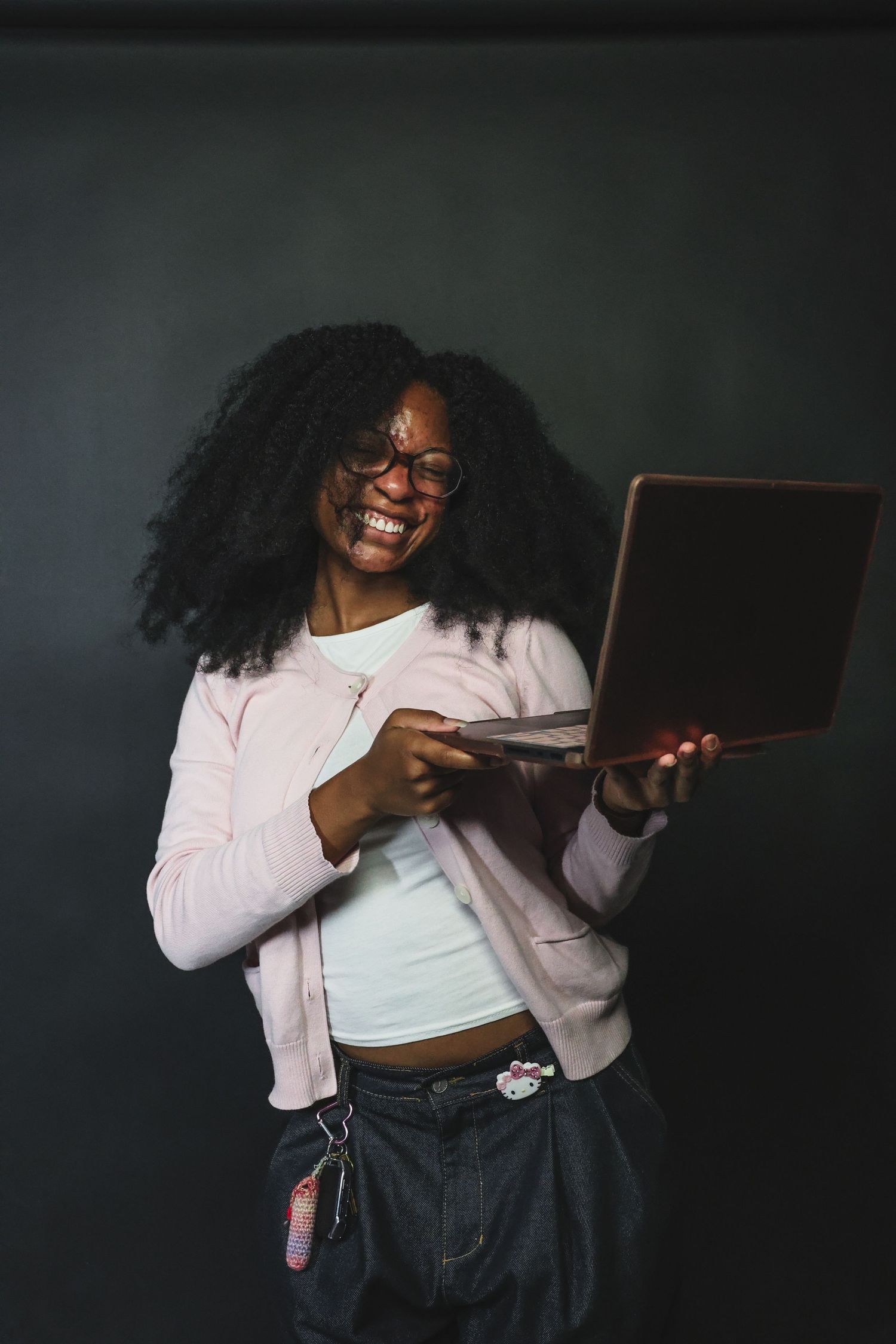 Happy Black woman with curly hair and glasses holding laptop, wearing pink jacket and jeans.