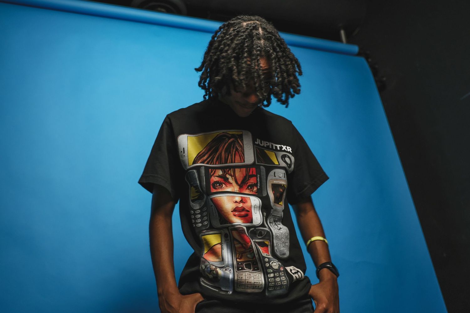 Young man with braids wearing a graphic black t-shirt poses against a blue backdrop.