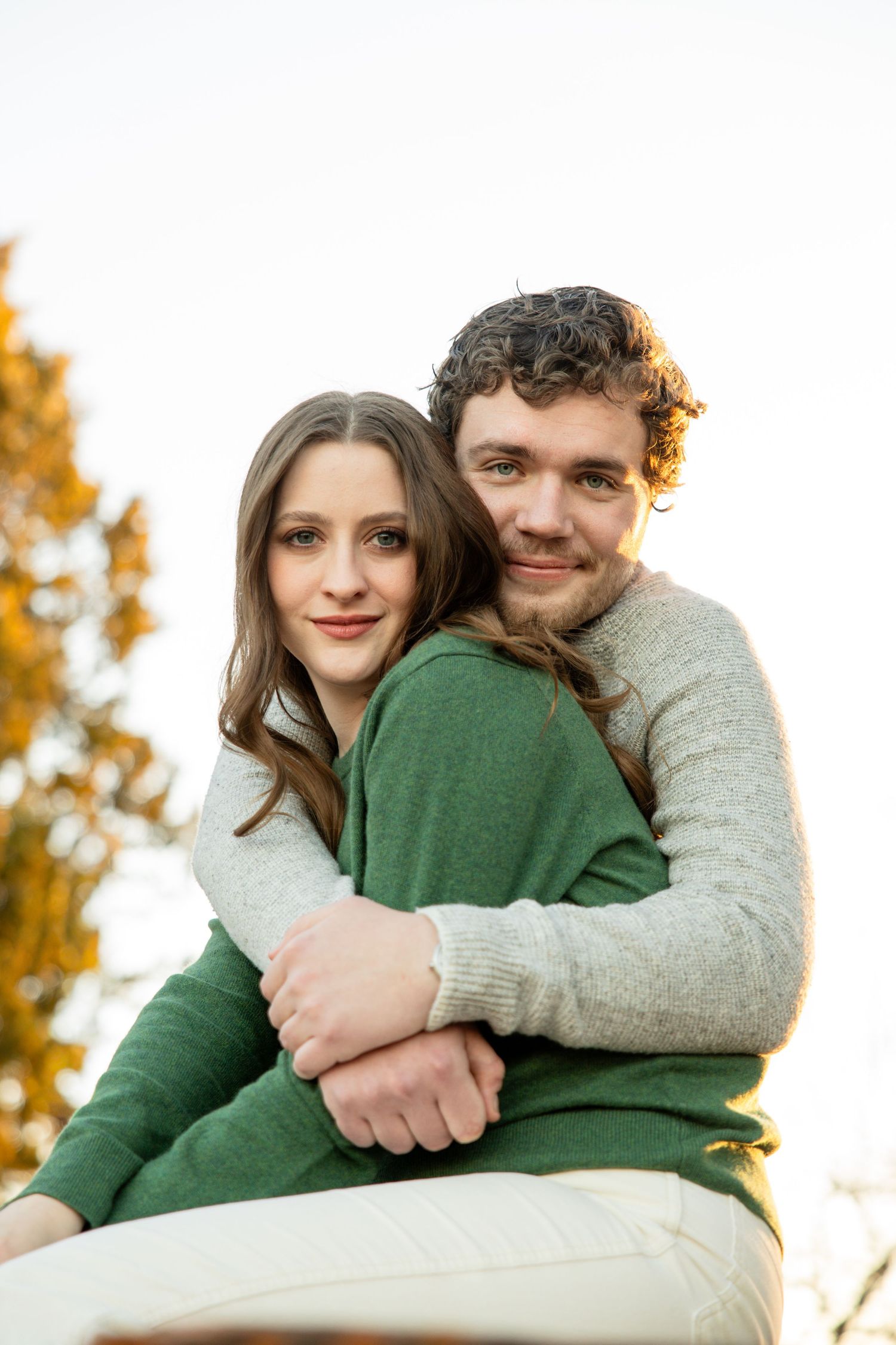 Man in gray sweater embracing woman in green sweater from behind during autumn outdoor photo session.