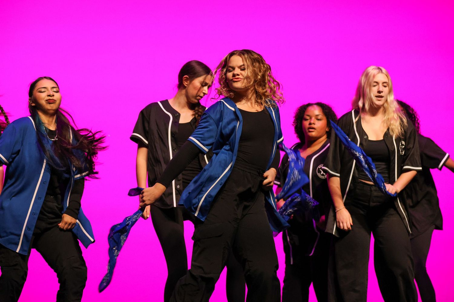 Group of women performing an energetic dance routine on stage with a vibrant pink background.