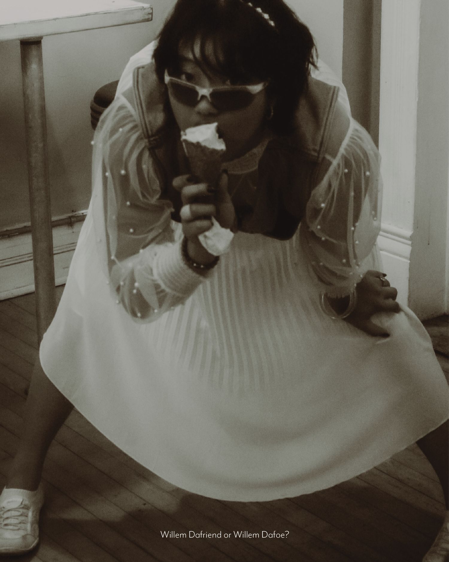 Person in white pleated dress and sunglasses crouching on wooden floor in sepia-toned photo.