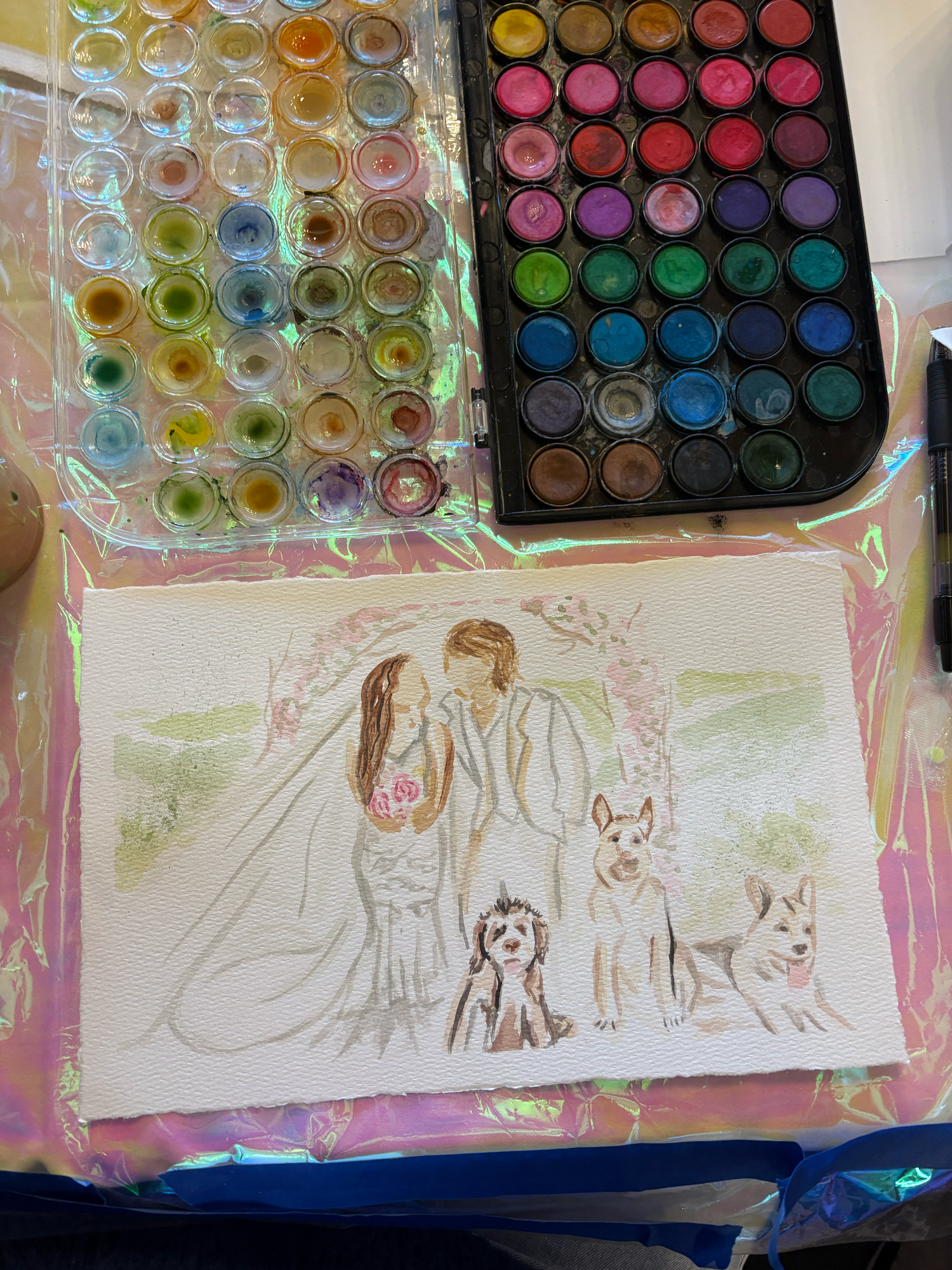 Watercolor wedding portrait painting of couple with dogs, surrounded by paint palettes on iridescent surface.