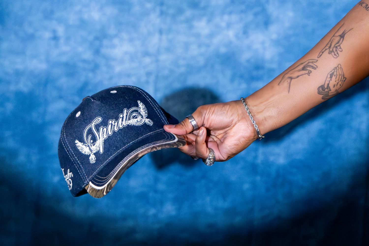Tattooed hand holding a navy Spirit embroidered baseball cap against blue textured background.