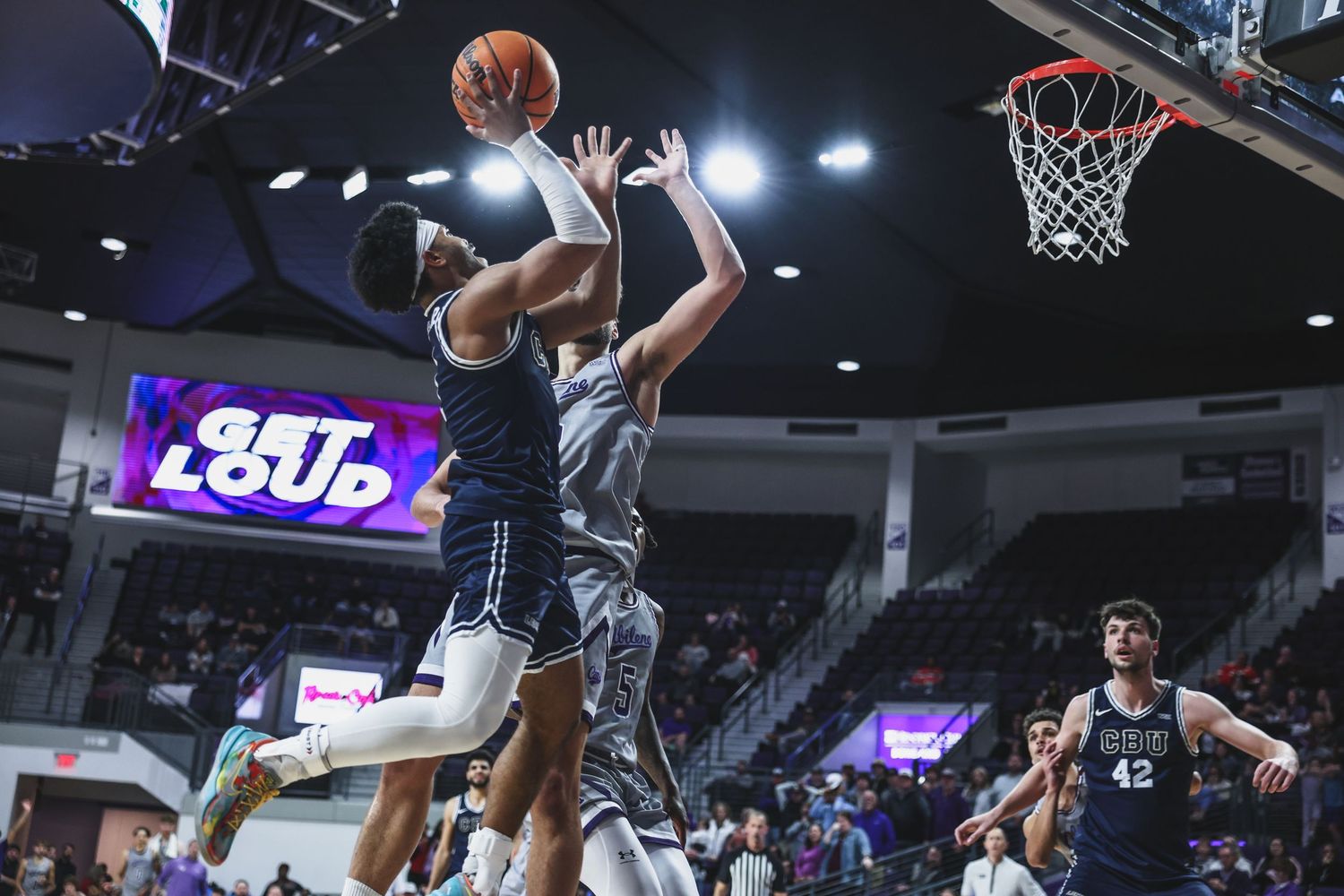 Basketball player drives to the basket for a layup against a defender in a college arena.