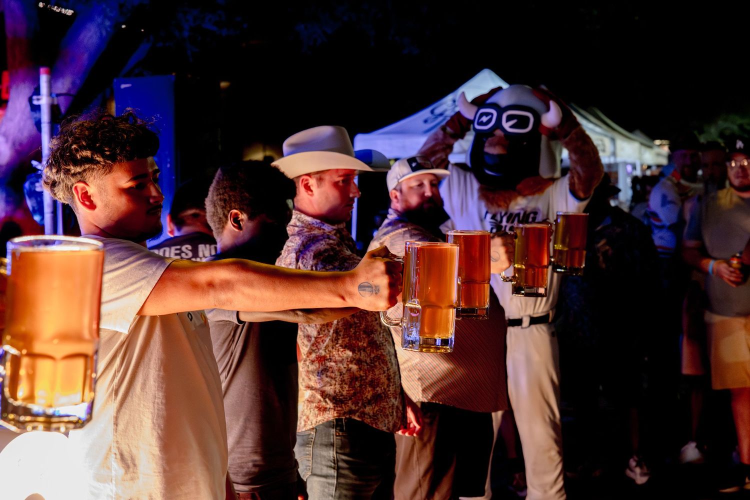 Group of men toasting with large beer mugs at an outdoor night festival event.