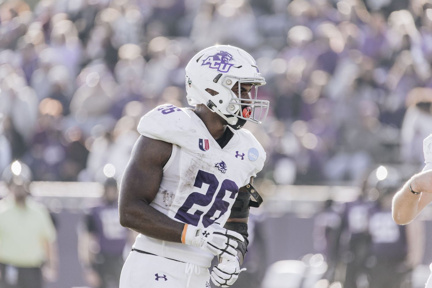 ACU football player #26 in white and purple uniform jogs on the field during a game.