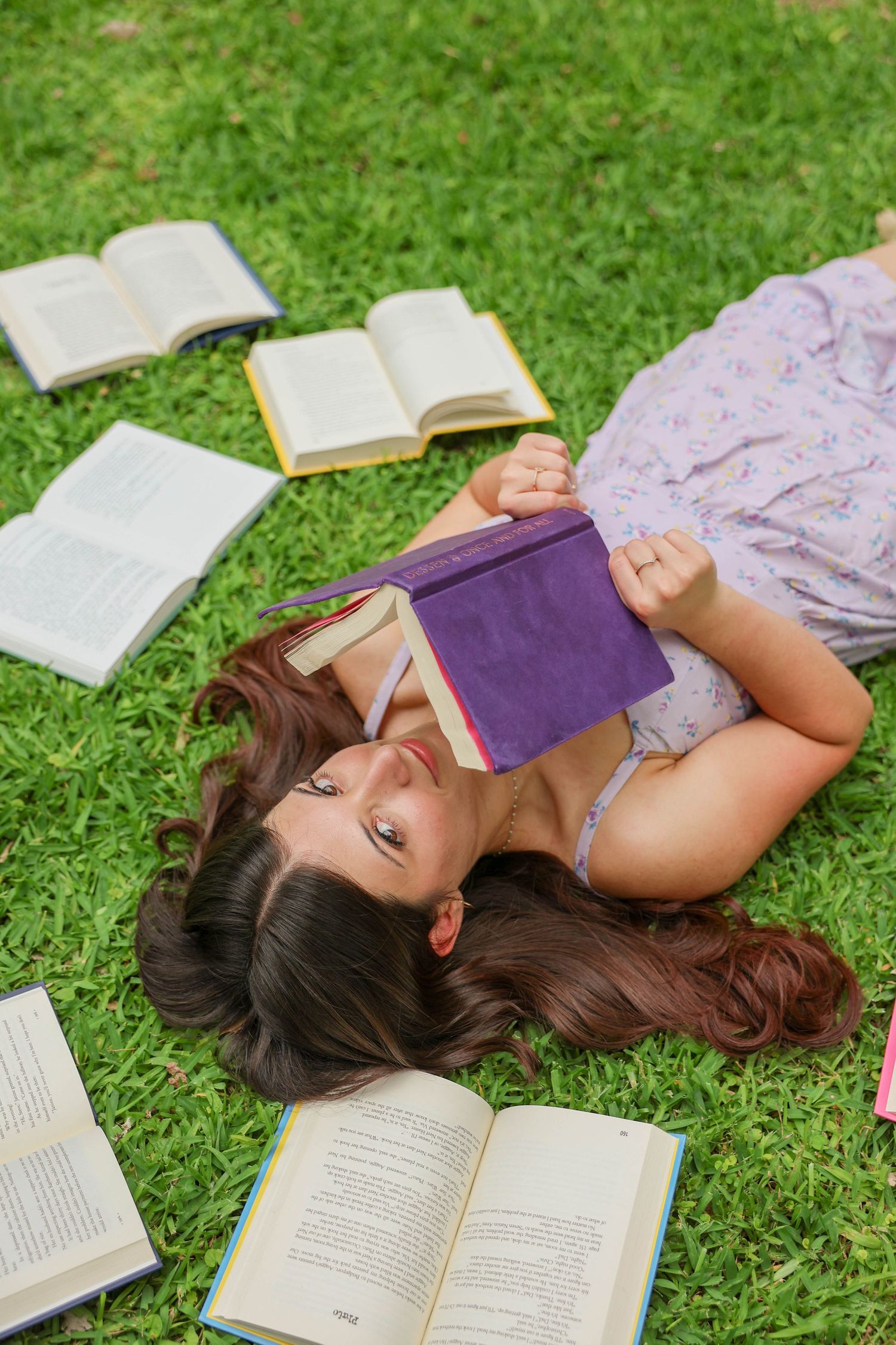 Young woman lying on green grass surrounded by open books, holding a purple book while relaxing outdoors.