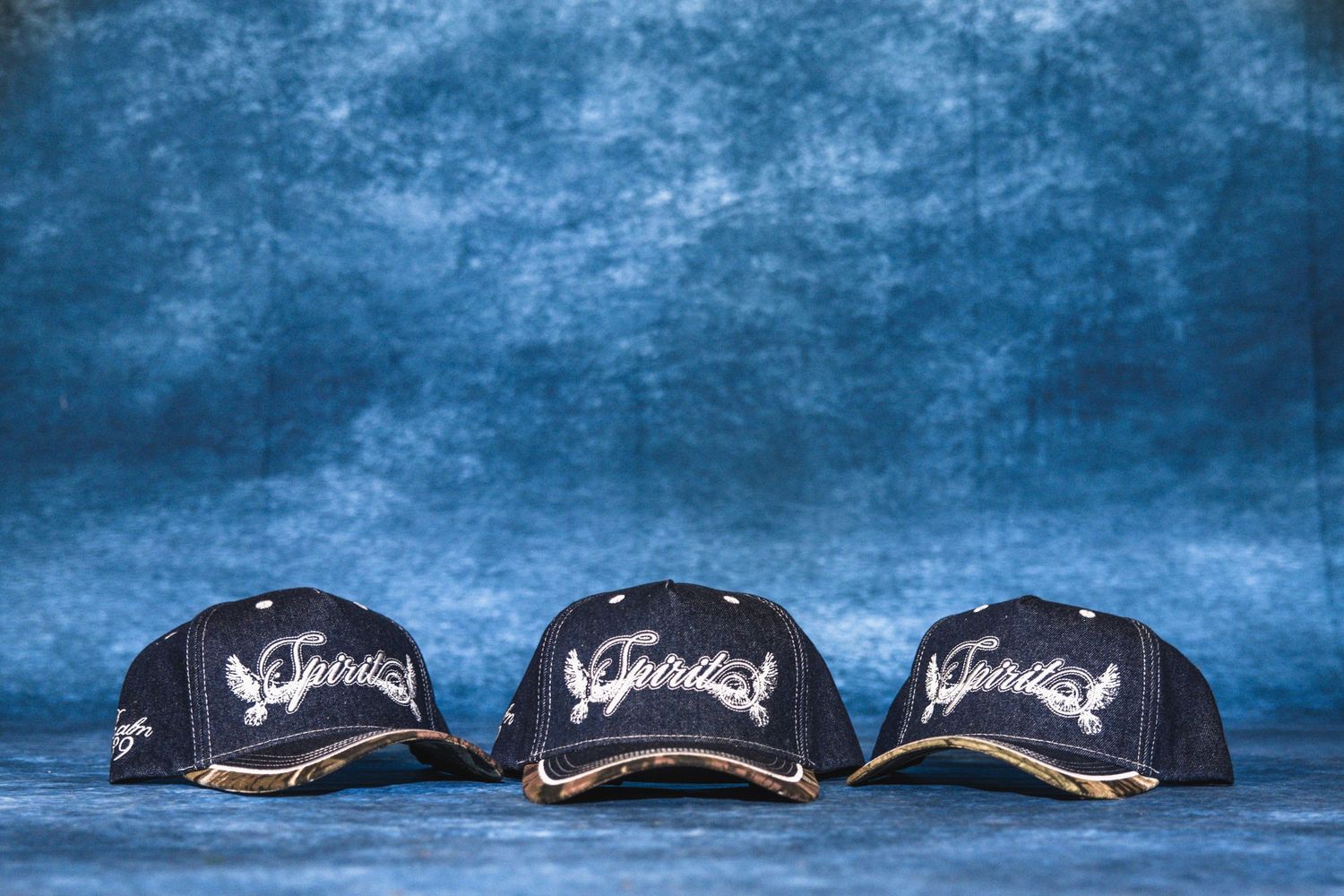 Three dark navy blue embroidered Spirit baseball caps displayed on a blue textured background.