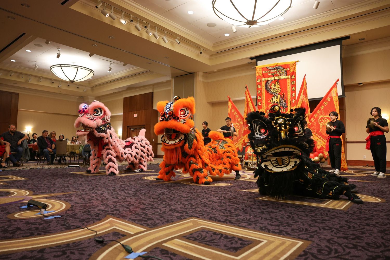 Three Chinese lion dancers in pink, orange, and black costumes perform in a ballroom with red banners.