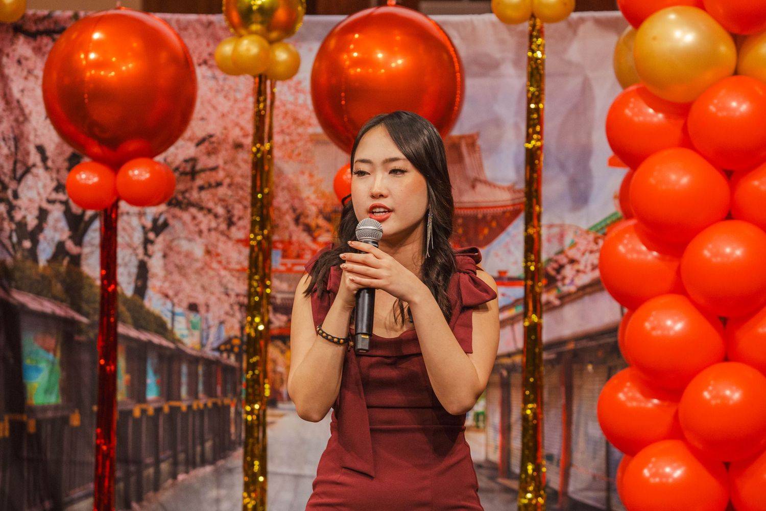 A young woman in a red qipao performs on stage surrounded by red and gold Lunar New Year balloon decor.