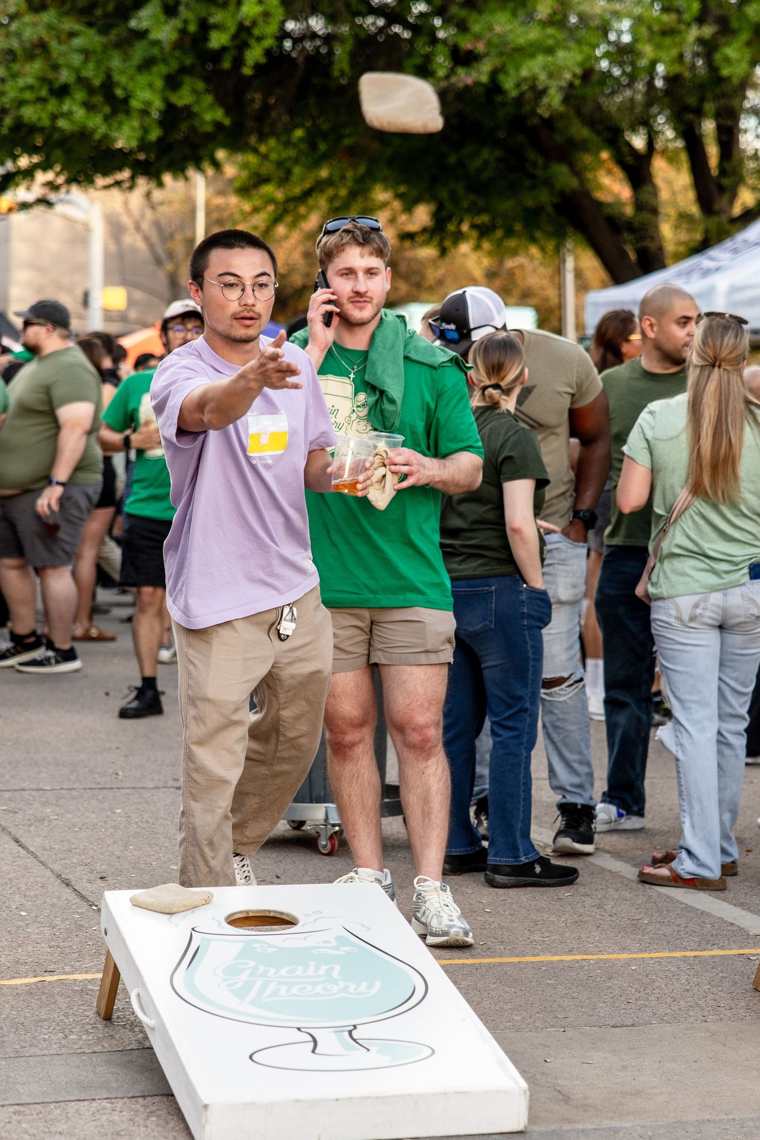Two men play cornhole at an outdoor festival, one pointing while holding a phone.