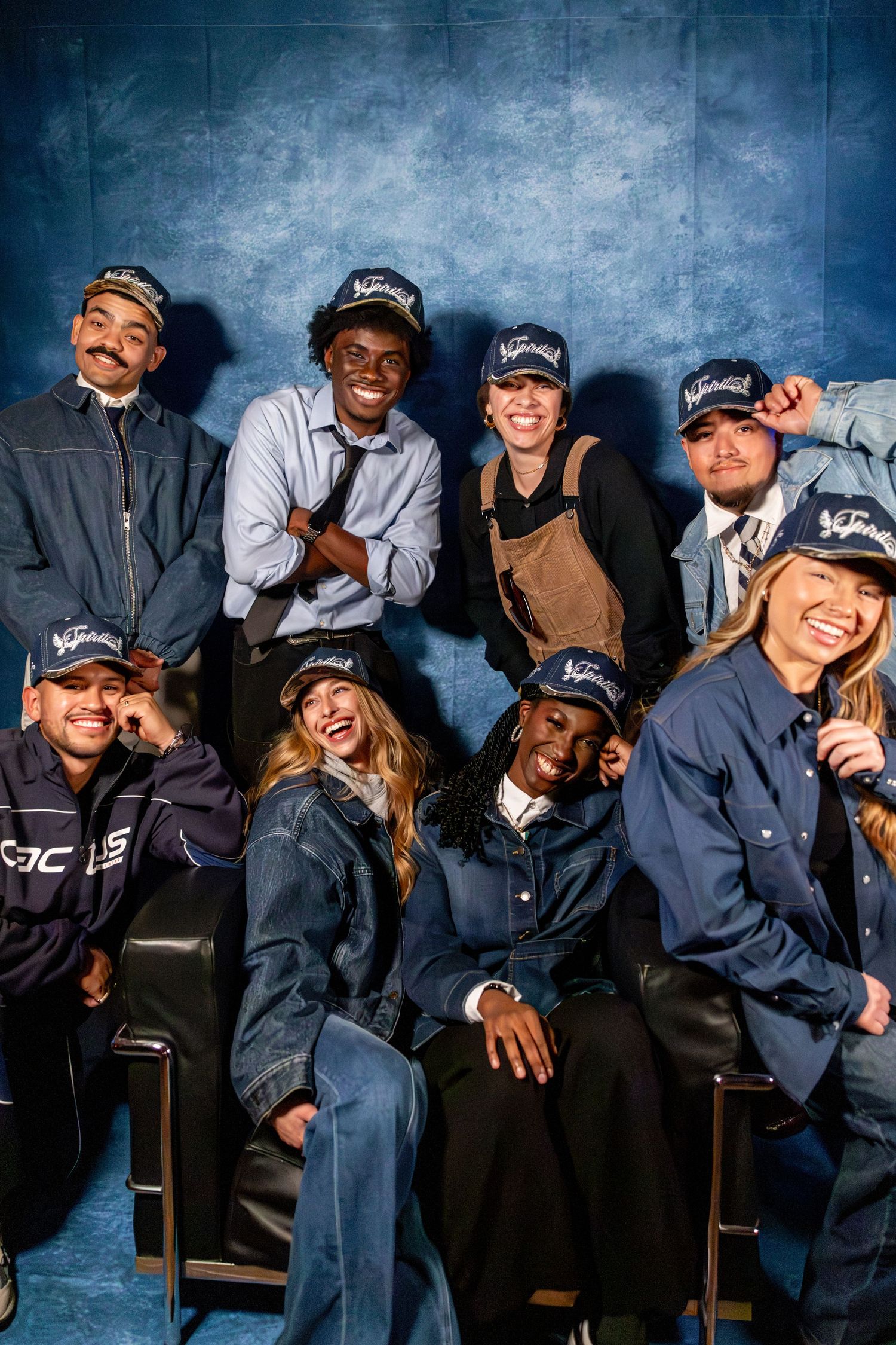Group of nine diverse friends pose together wearing matching navy branded caps indoors.